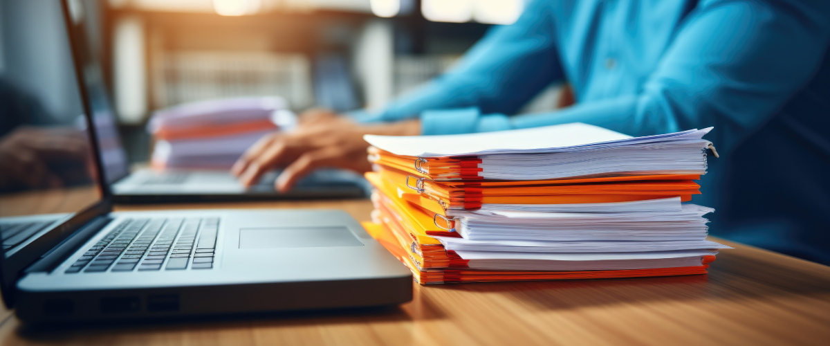 Close-up of a man's hands typing on a laptop keyboard, with a stack of paperwork beside them